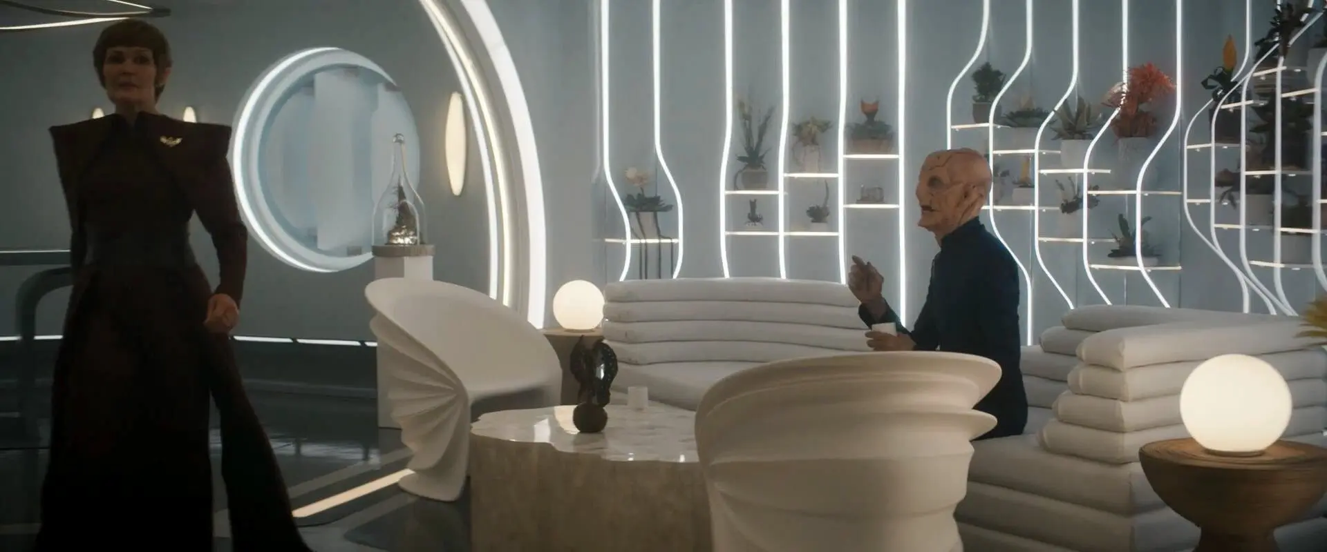 A screencap from Star Trek: Discovery, depicting Saru sitting on a white couch that resembles a lot of layers of white cushions, near white chairs which look somehow wrinkled.